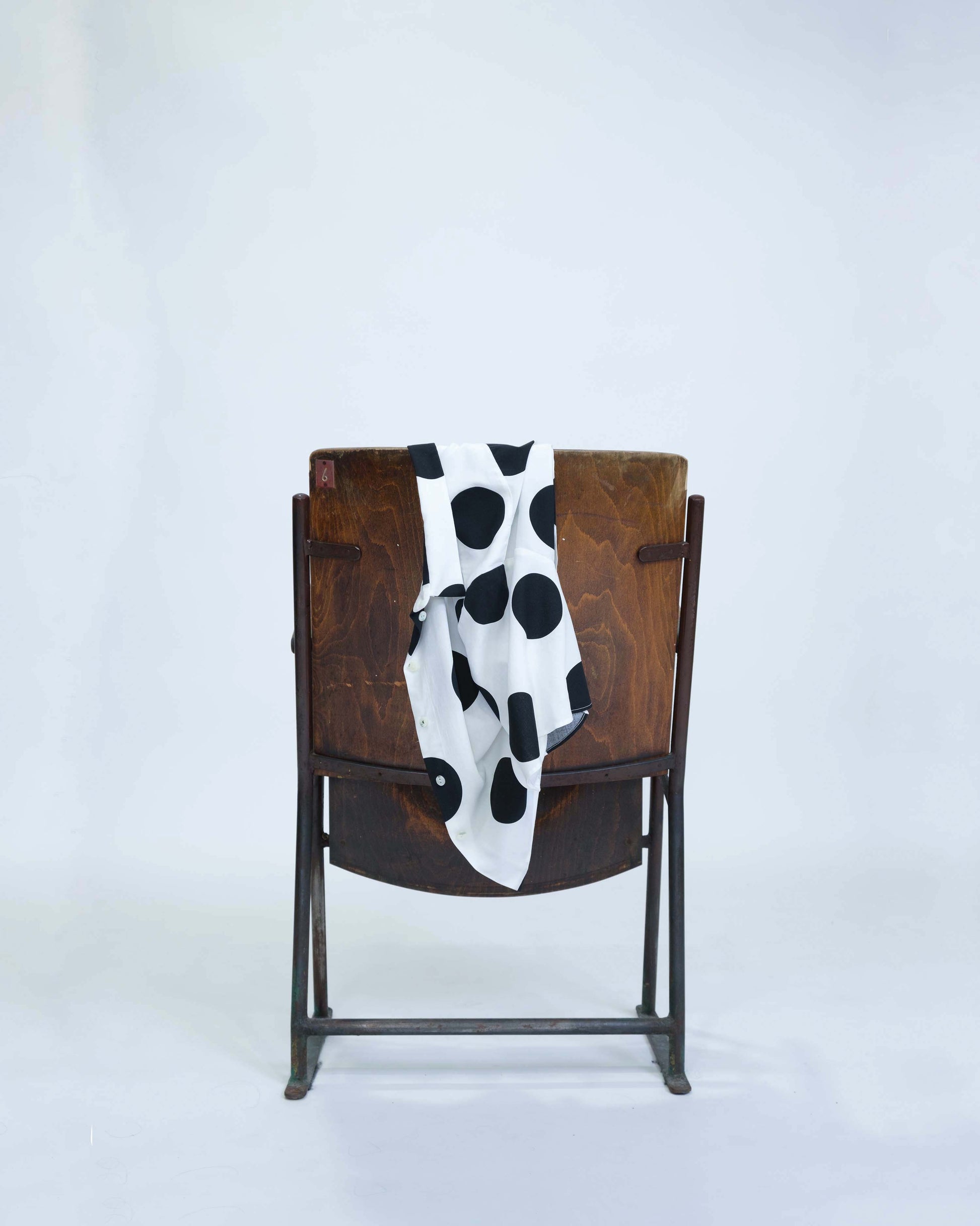Chemisette pour femme réalisée à la main en deux exemplaires, en viscose blanche à gros pois noir, posée sur le dossier d'une chaise en bois, détail tombé du tissu. 